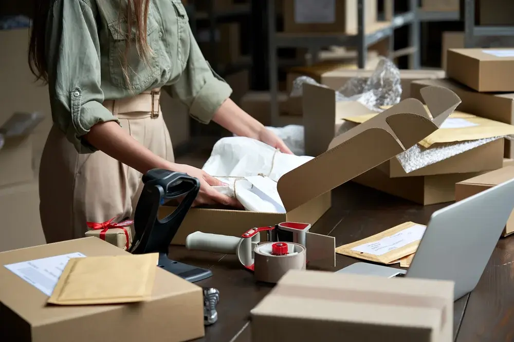 Shows a woman at a table, packing a shirt into a cardboard box, to be shipped as an online order.