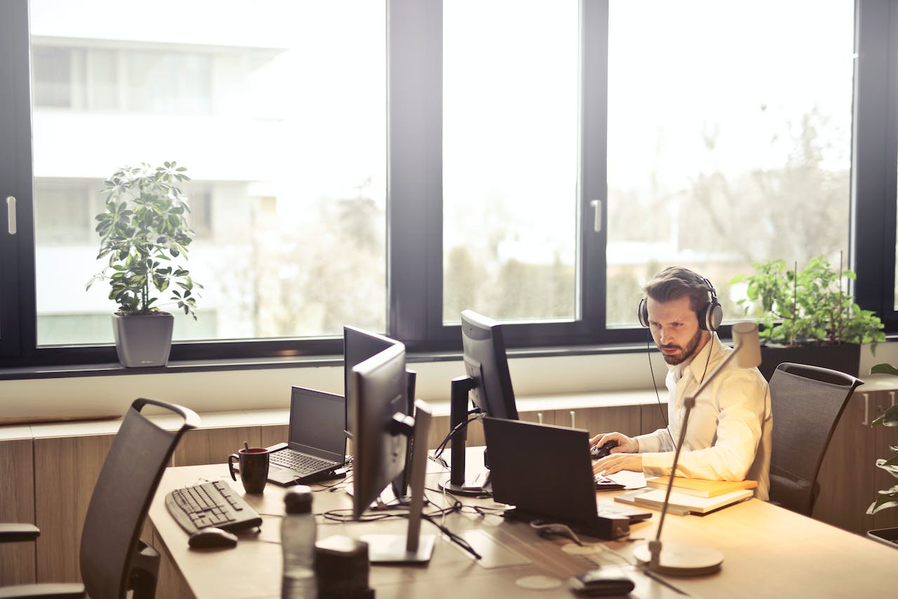 Shows a man with headphones on, at a workdesk by a large window, hard at work catering to customer needs over call.