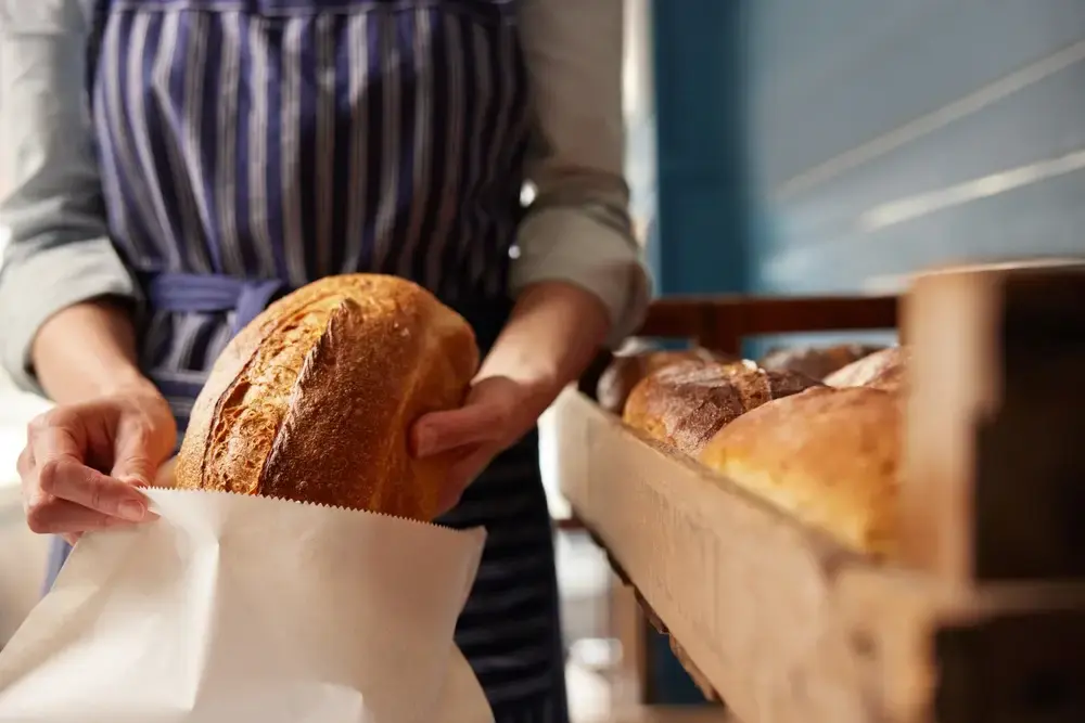 A baker is packing loaves of bread in eco-friendly paper packaging.