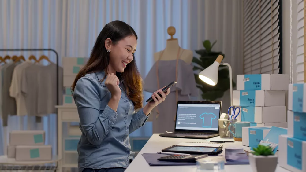 A woman in her retail business excitedly using her laptop and phone for a successful mobile eCommerce sale.