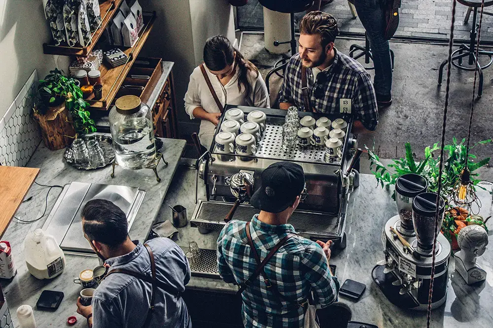 Top view of a cafe counter's daily operations, with staff talking to customers as they prepare drinks.