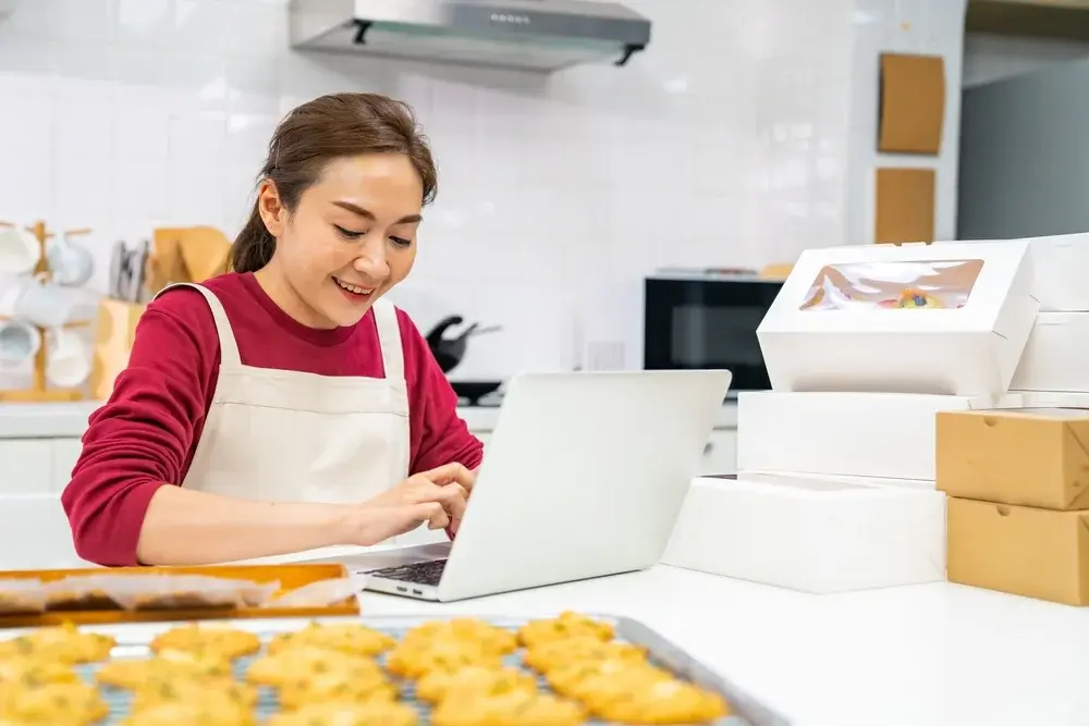 Shows a woman in the kitchen, working on a laptop, happily managing her baking online food business.